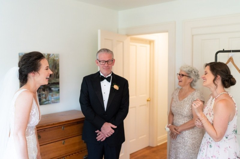 Bride, father, and two women sharing a joyful moment in a bedroom during a wedding celebration.