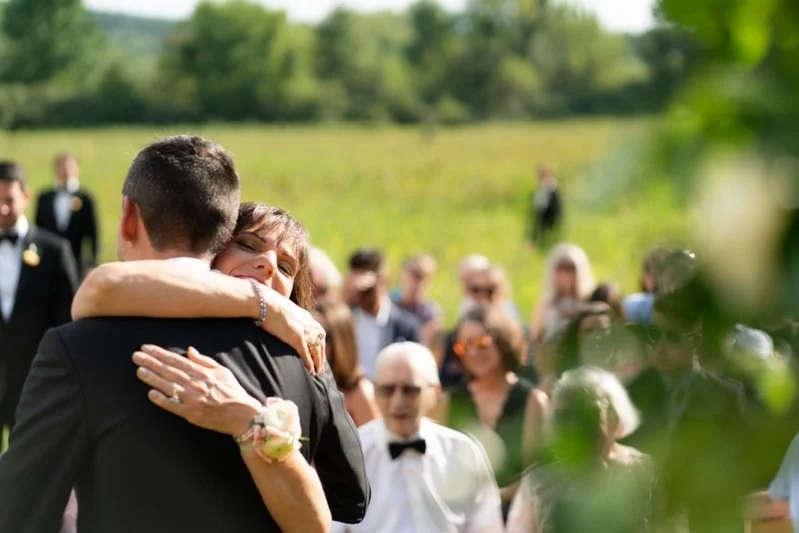 A woman embracing a man outdoors at a wedding, with guests in the background.
