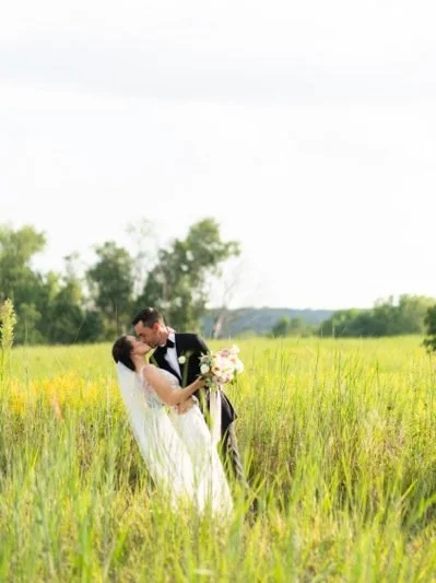 A bride and groom standing in a grassy field, kissing, with the groom holding a bouquet of flowers.