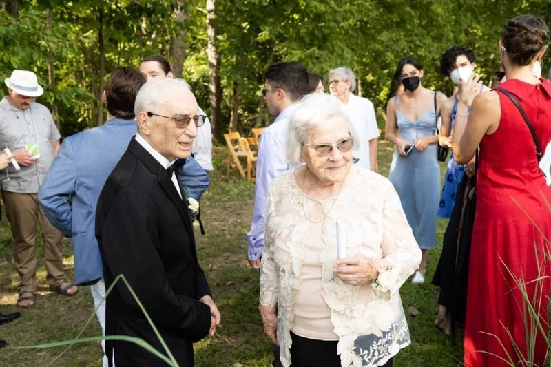 An outdoor social gathering with elderly and younger people, some wearing masks, among trees, with chairs in the background.