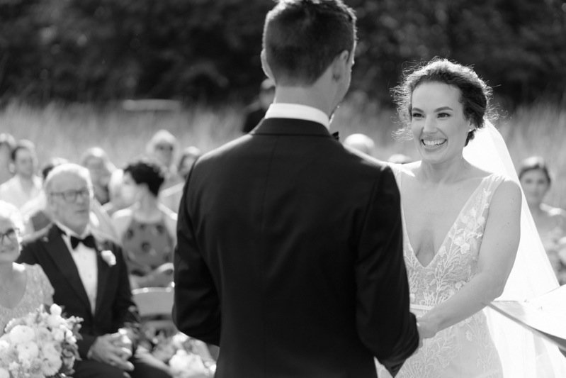 A wedding ceremony outdoors with a bride and groom facing each other, surrounded by seated guests.