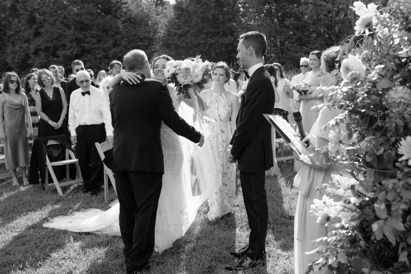 A wedding ceremony outdoors with the bride and groom exchanging vows while the officiant holds a book. Guests watch in the background, and the bride is holding a large bouquet of flowers.
