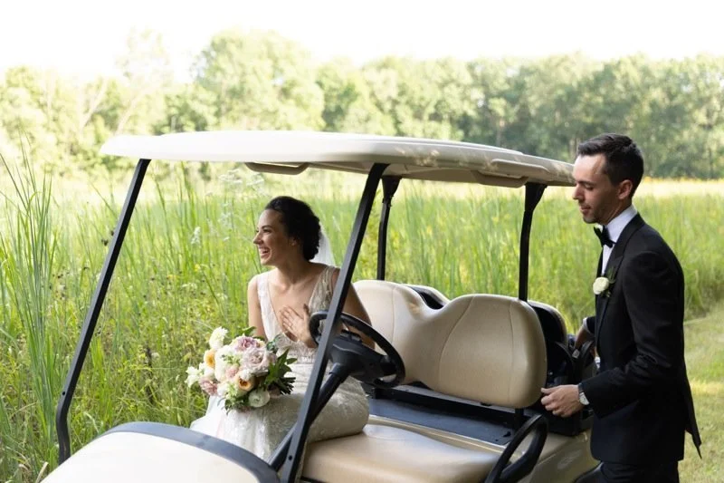 Bride sitting in a golf cart holding a bouquet while smiling, groom standing next to the cart in a tuxedo, outdoors in a green field with trees in the background, daytime.