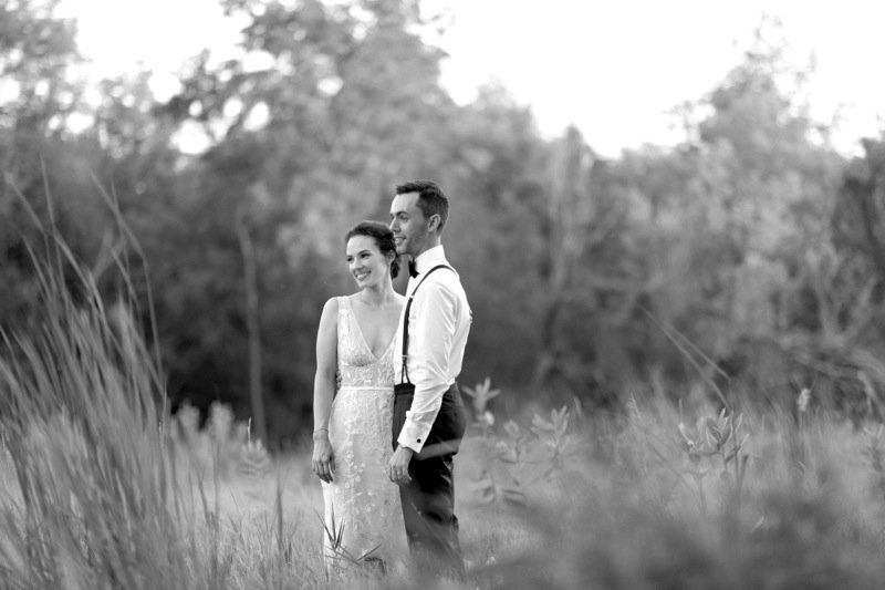 A couple in wedding attire standing in a field, smiling with trees in the background.