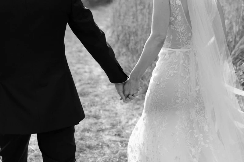 A bride and groom holding hands outdoors, with the bride wearing a wedding dress and veil, and the groom in a dark suit.