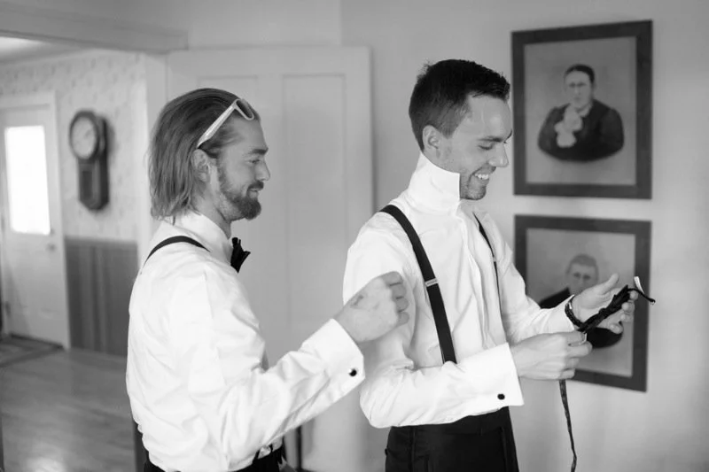 Two men dressed in formal attire, one with suspenders and the other with a bowtie, are smiling and looking at a smartphone indoors with framed portraits on the wall.