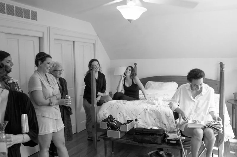 Group of women gathered in a bedroom, some sitting on the bed, laughing and chatting, while one woman looks at a book or magazine.