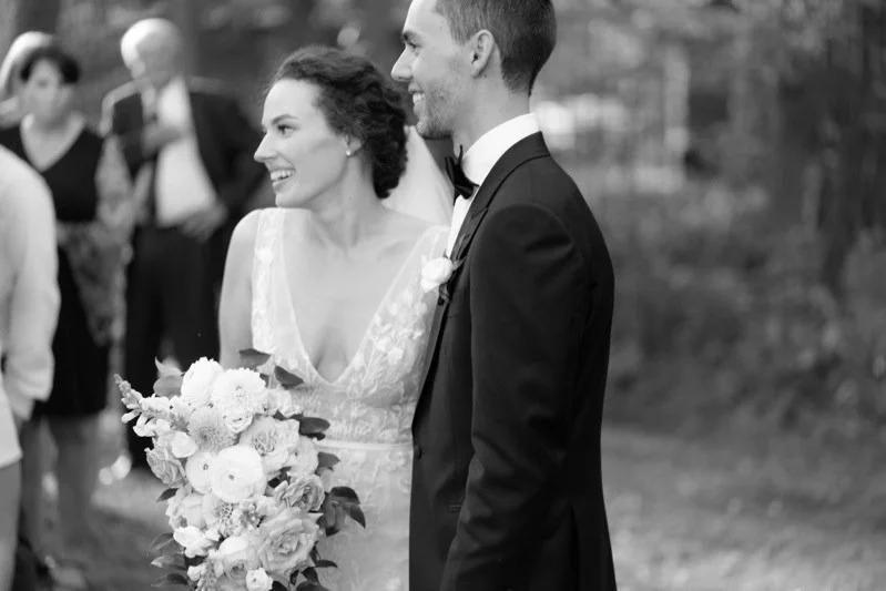 A black and white photo of a bride and groom smiling at each other during their wedding ceremony. The bride holds a bouquet of flowers, and the groom is dressed in a tuxedo with a bow tie.