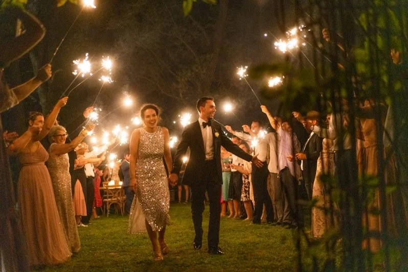 Couple in formal attire walking hand in hand through guests holding sparklers at night during a celebration or wedding reception outdoors.