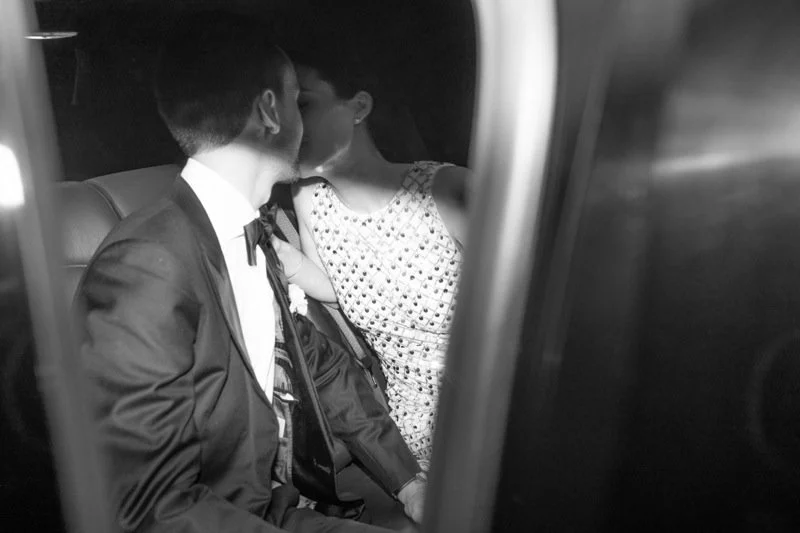A black and white photo of a couple kissing inside a vehicle, with the man in a suit and the woman in a patterned dress.