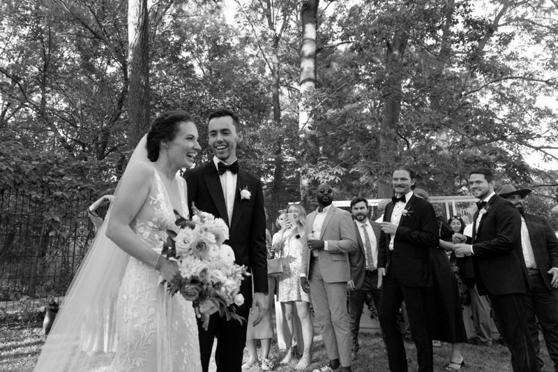 A black-and-white photo of a bride and groom smiling with guests at an outdoor wedding reception with trees in the background.