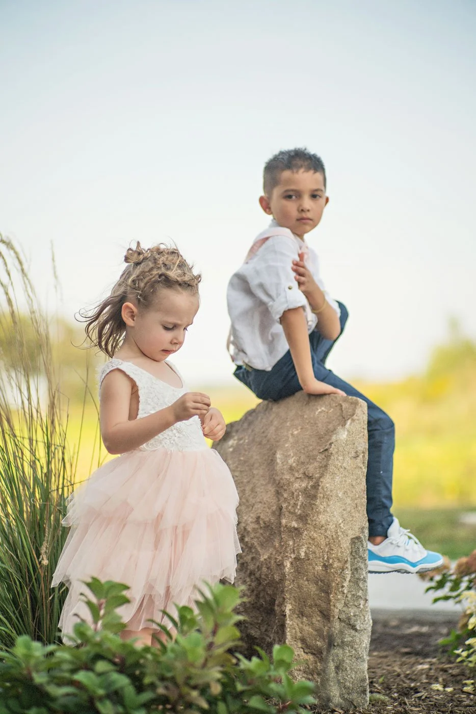 A young girl in a light pink dress with a lace top and tulle skirt stands next to a boy sitting on a large rock. The boy wears a white long-sleeve shirt, dark pants, and sneakers. They are outdoors near greenery and appear to be in a park or garden s