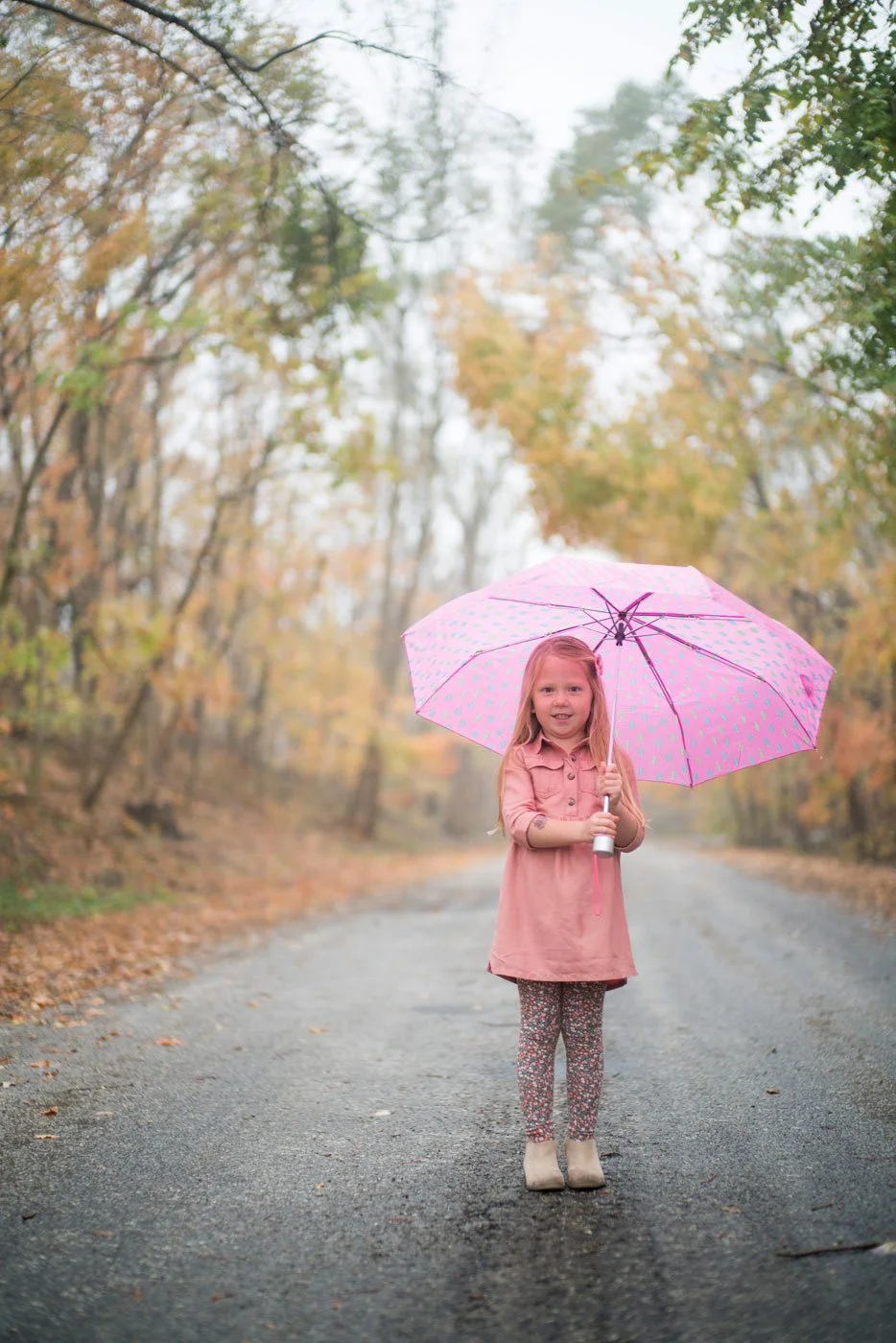 A young girl with long blonde hair wearing a pink coat and patterned pants stands on a gravel road holding a pink umbrella with teal dots. The background has autumn trees with yellow and orange leaves.