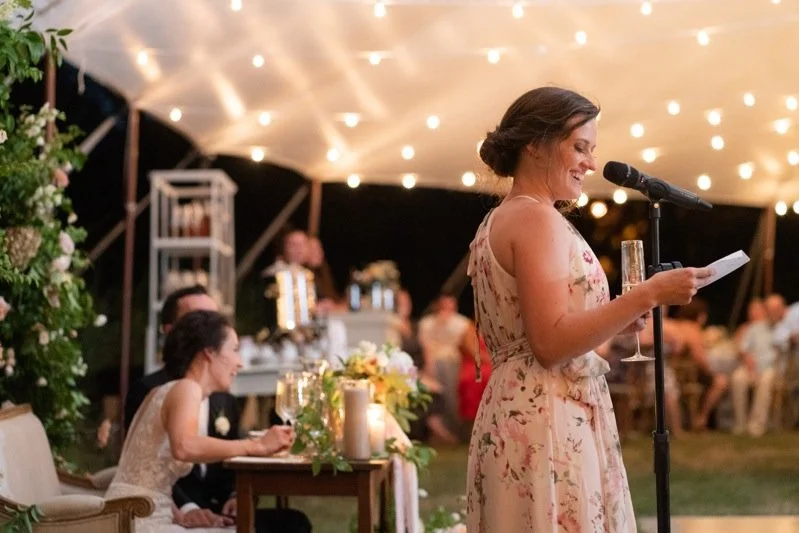 A woman giving a speech at a wedding reception, standing at a microphone holding a piece of paper, surrounded by guests in a decorated outdoor tent.