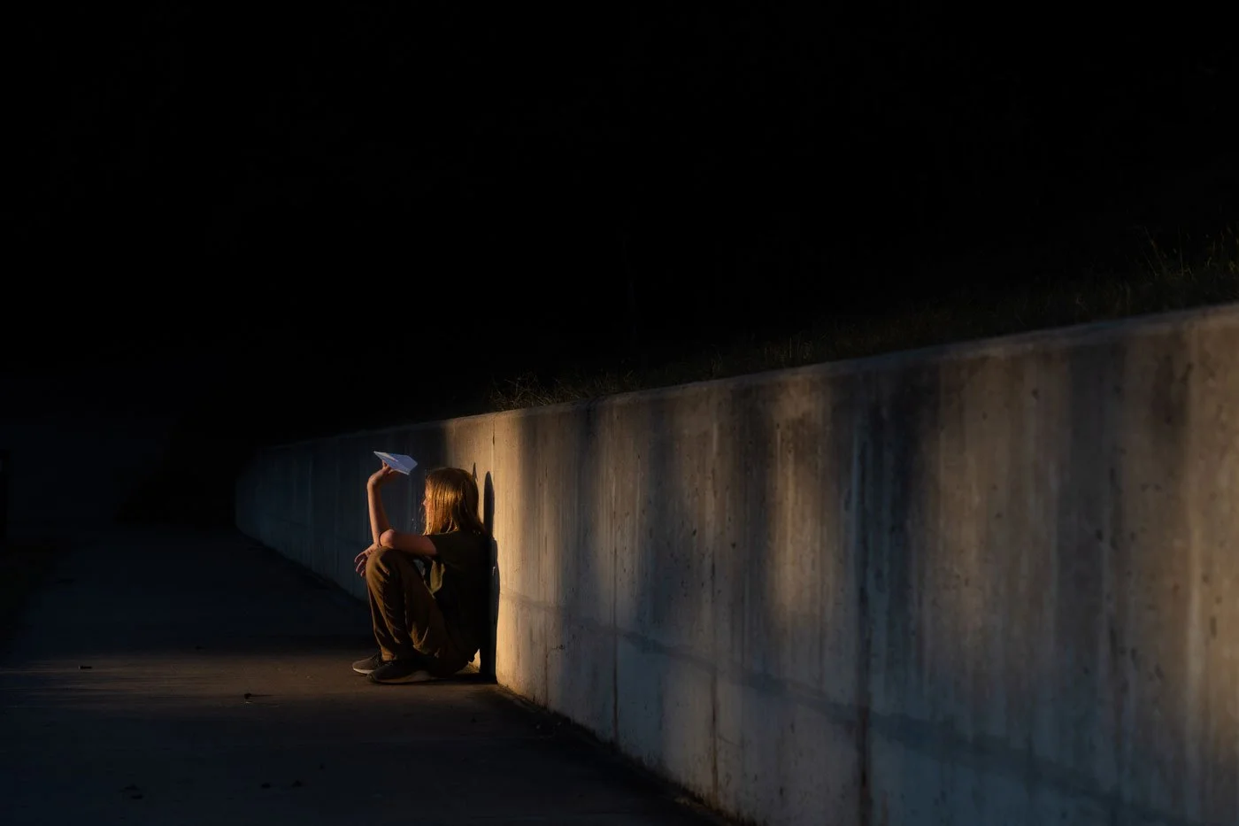 A girl sitting on the ground next to a concrete wall at night, holding a paper plane and looking away.