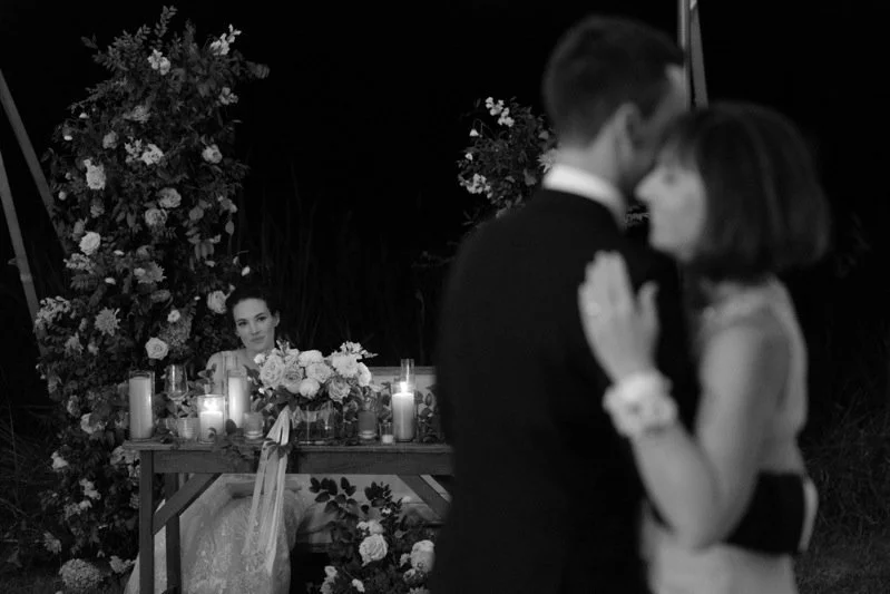 A black and white photo of a wedding reception, with a couple dancing in the foreground and a woman sitting at a decorated table in the background.