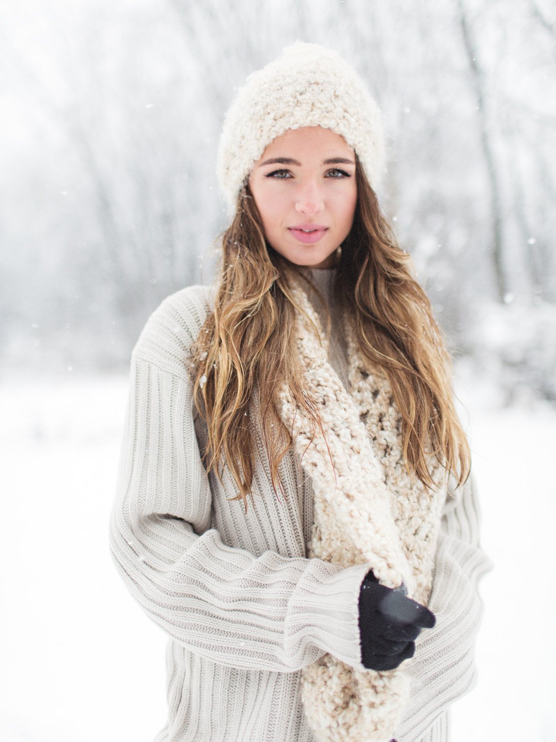 A young woman with long wavy hair wearing a cream knit hat, scarf, a beige sweater, and black gloves standing outdoors in a snowy winter landscape.
