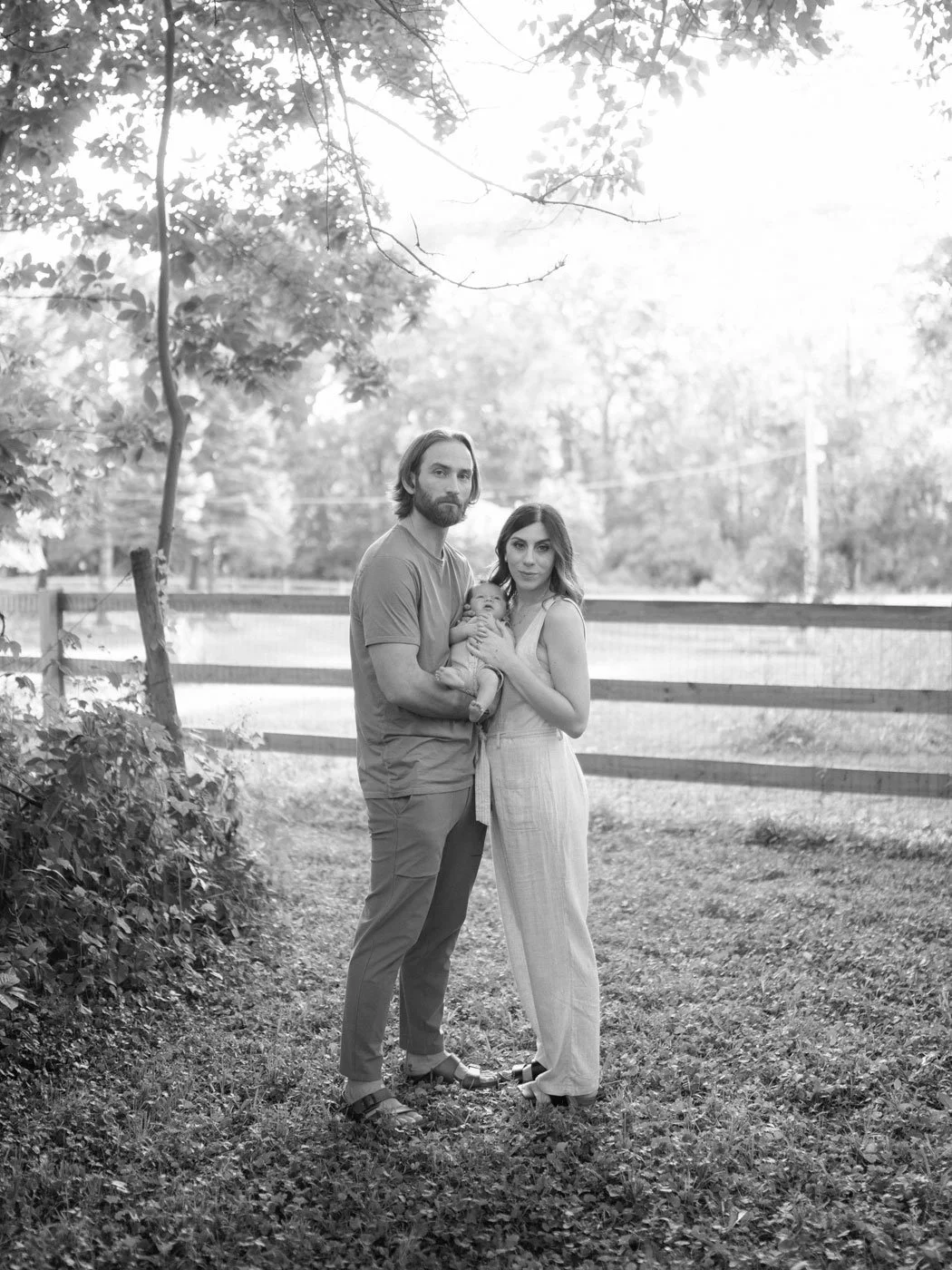A family of three, a man, woman, and baby, standing outdoors in a grassy area with trees and a fence in the background.