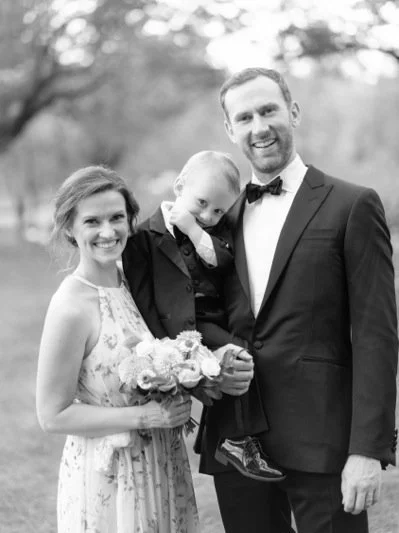 A black and white photo of a happy family outdoors, with a woman holding a bouquet, a young boy, and a man in a suit and bowtie.
