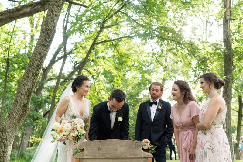 A bride and groom at their wedding ceremony outdoors, signing a document surrounded by three women and one man, with trees and sunlight overhead.