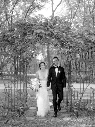 A bride and groom walking through a garden archway on their wedding day, holding hands and smiling.