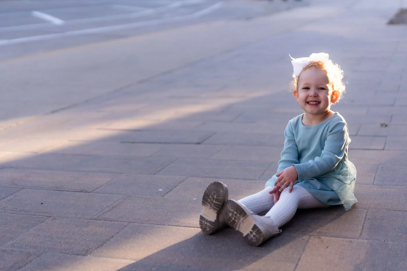 A young girl with curly blonde hair, wearing a light blue dress, white tights, and boots, sitting on the sidewalk and smiling at the camera.