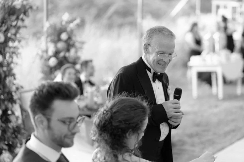 A man in a tuxedo with a microphone, smiling at a wedding reception.