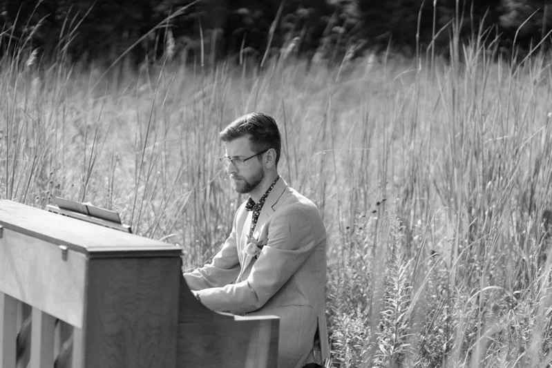 A man wearing glasses and a light-colored suit playing a piano outdoors in a grassy field.