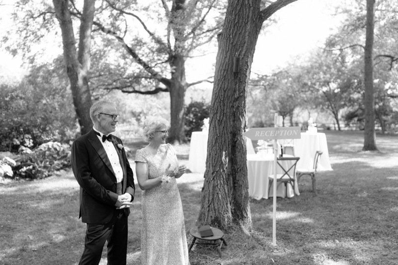 A man and woman are standing outdoors near a tree, dressed in formal attire for a wedding or outdoor event, with a reception table and sign in the background.