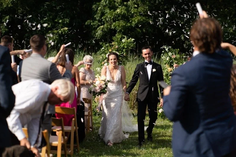 Bride and groom walking hand in hand on a grassy outdoor wedding aisle, surrounded by guests cheering and taking photos, with trees and greenery in the background.