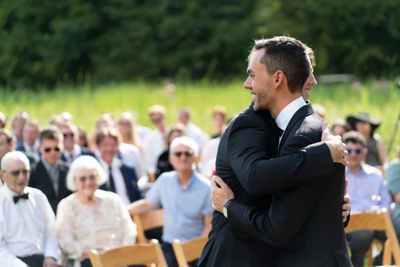 Two men in suits hug at an outdoor wedding ceremony with seated guests watching.