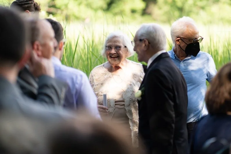 Elderly woman smiling and laughing at a gathering outdoors on a sunny day, surrounded by other people, some wearing masks.