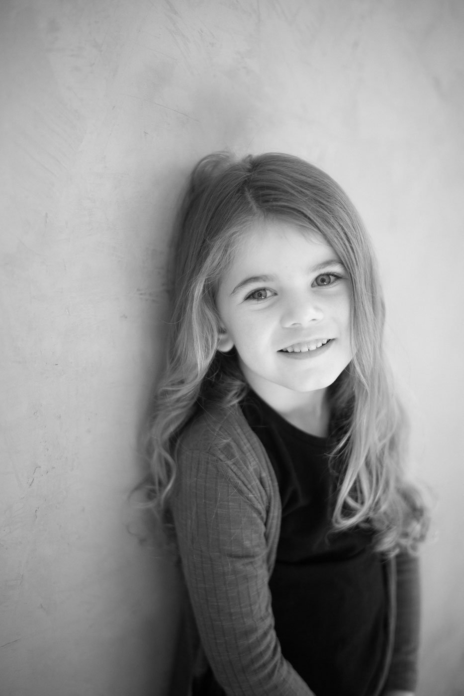 Black and white photo of a young girl with long wavy hair smiling, leaning against a textured wall.