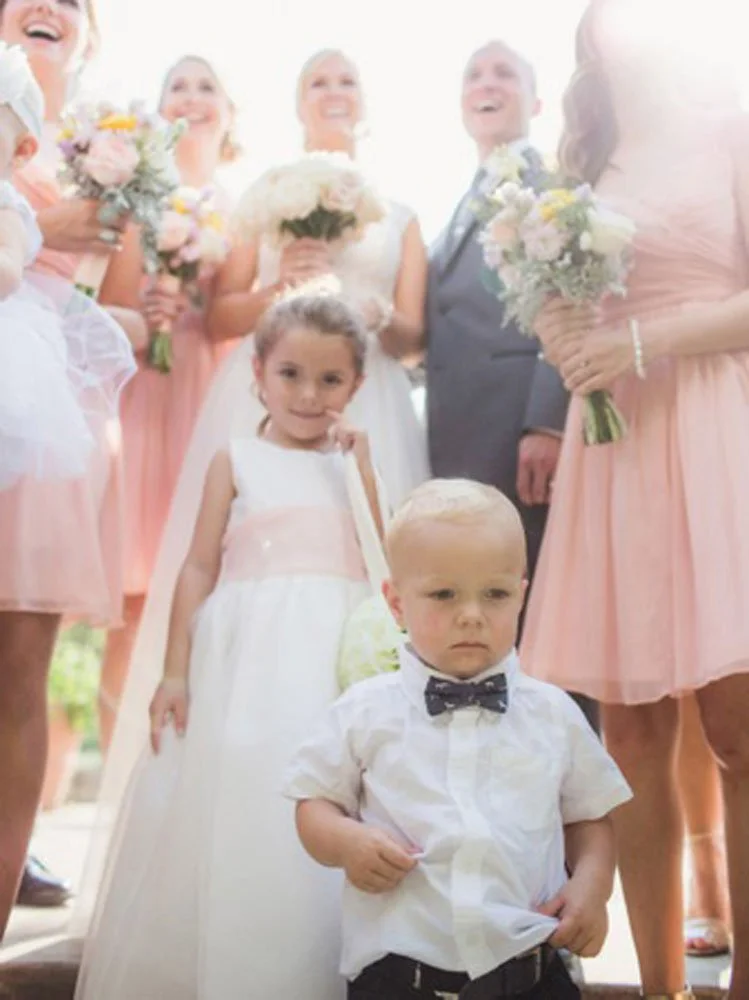 Children and adults in formal wedding attire, with a group of women in pink dresses and men in suits, including a young girl in a white dress and boy in a white shirt with a bow tie, at an outdoor wedding celebration.