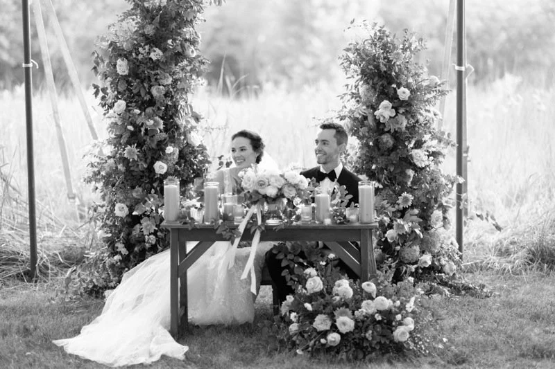 A bride and groom sitting at a decorated table during their wedding reception, surrounded by floral arrangements and candles outdoors.