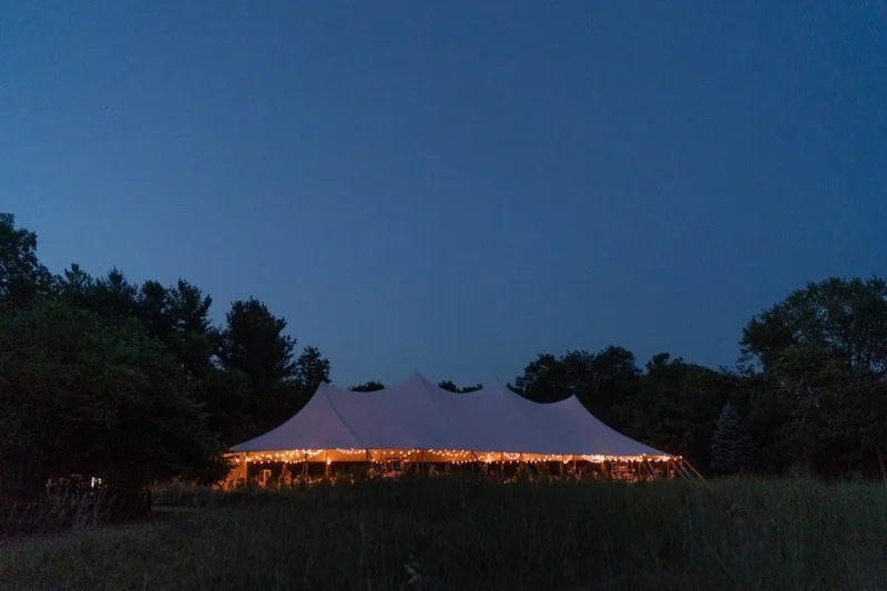 A large white tent with decorative peaks, illuminated with string lights at night, set in a grassy field surrounded by trees.