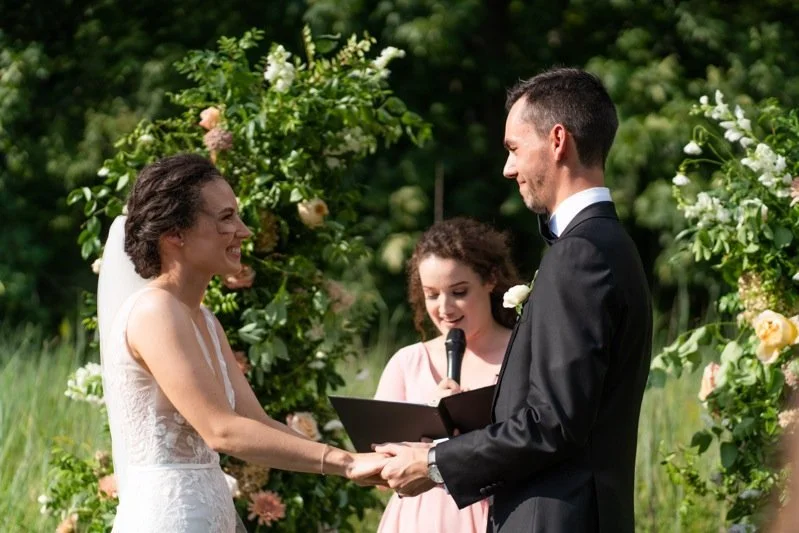 A bride and groom holding hands during an outdoor wedding ceremony, with an officiant reading from a book, surrounded by floral arrangements.