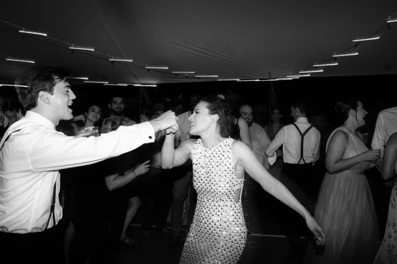 A black and white photo of a couple dancing and holding hands at a lively social event, surrounded by other dancing guests.