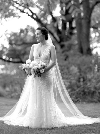 A bride in a white wedding dress holding a bouquet of flowers outdoors, smiling, with trees in the background.