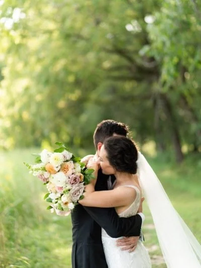 A bride and groom embracing outdoors, the bride holding a bouquet of flowers.