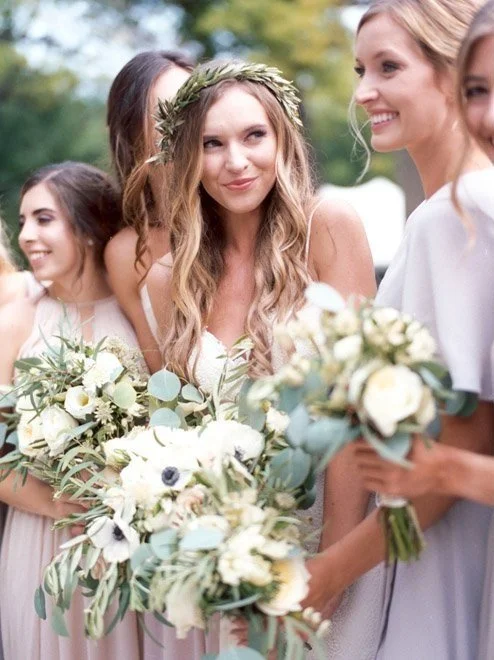 A woman with long wavy hair and a floral crown holding a bouquet of white flowers, standing with three women dressed in pale-colored dresses, outdoors during daytime.