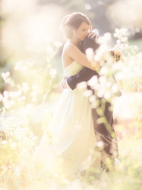 Bride and groom embracing in a sunlit outdoor garden, surrounded by blooming flowers.