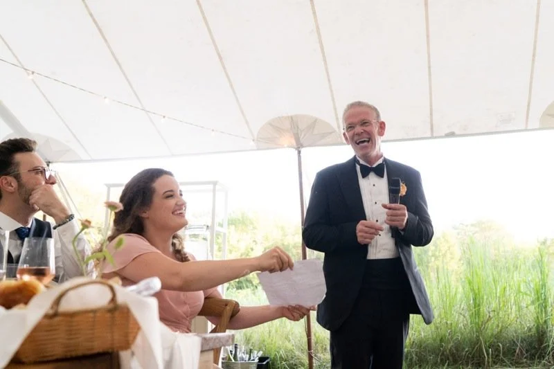 A wedding reception with a man in a tuxedo giving a toast as a woman and a man, both seated at the table, smile and listen.