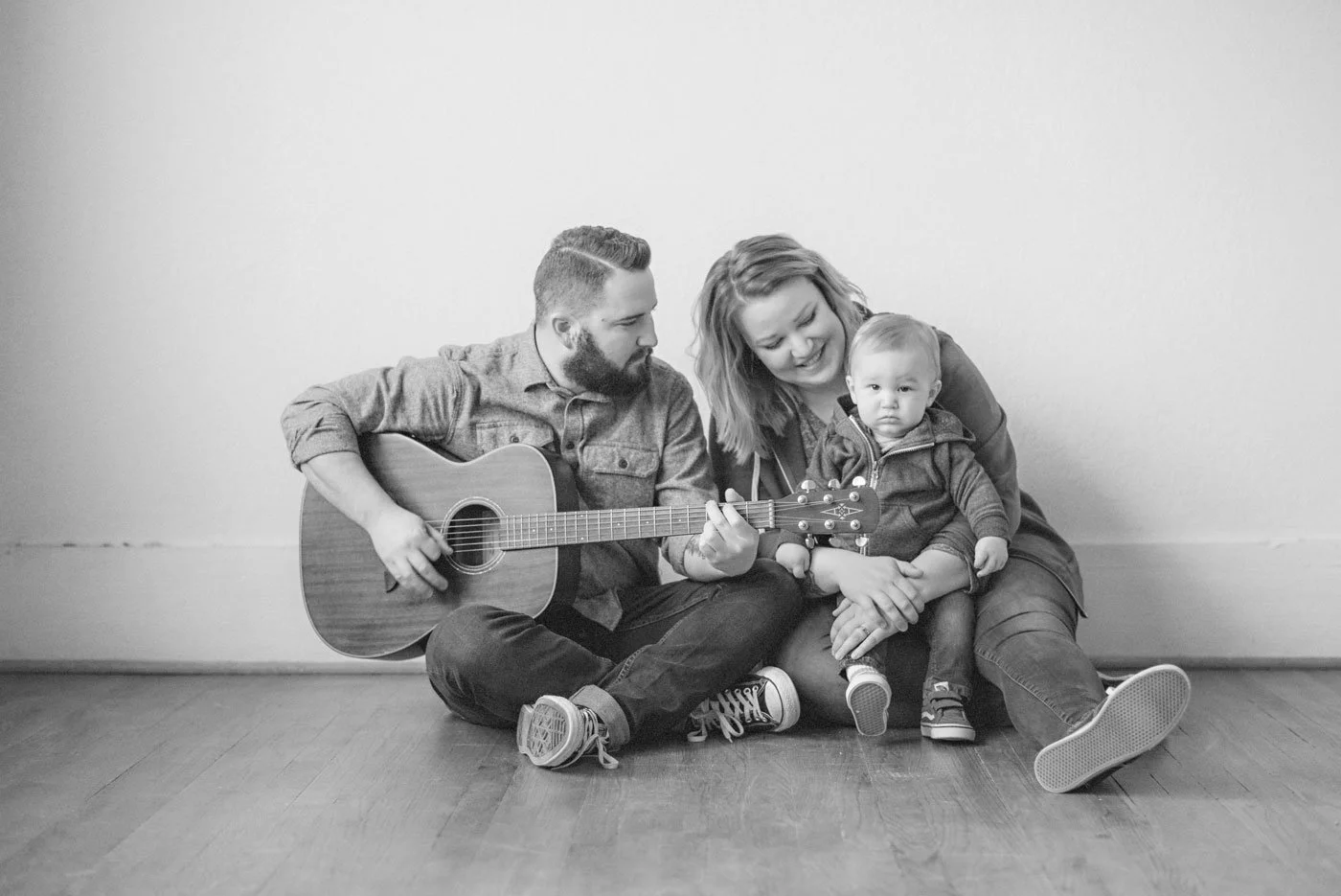 A black and white photo of a family of three sitting on the floor against a plain wall. The man is playing guitar, the woman is smiling and holding their young child, who looks curious.