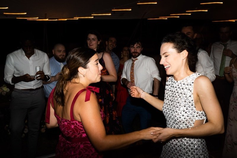 Two women dancing together at a party, smiling and holding hands, with other guests watching in the background.