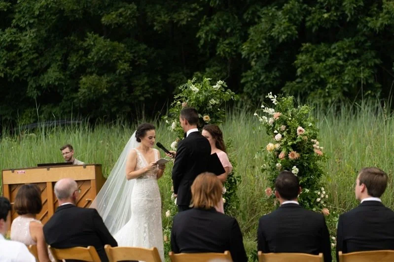 Bride and groom exchanging vows outdoors at a wedding ceremony with guests seated, a musician playing a keyboard in the background, and decorated floral arch.