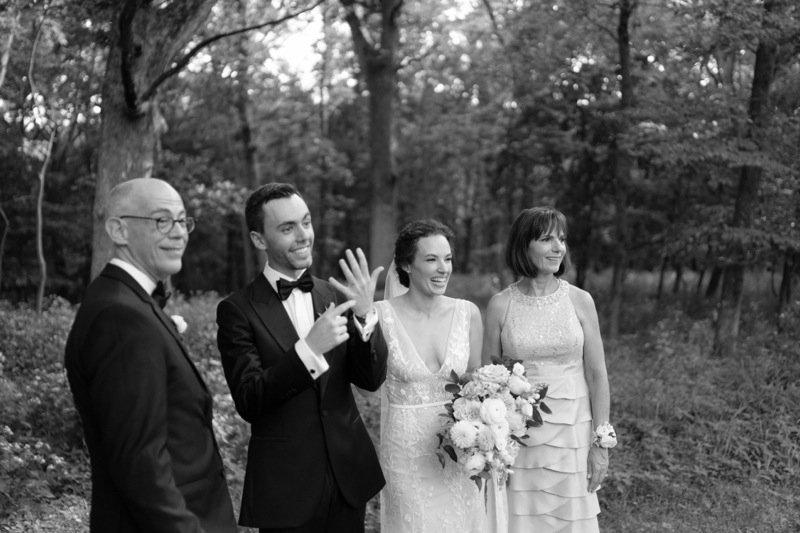 Black and white photo of a wedding party outdoors in a wooded area, featuring a groom, bride, and two other women, all dressed in formal attire.