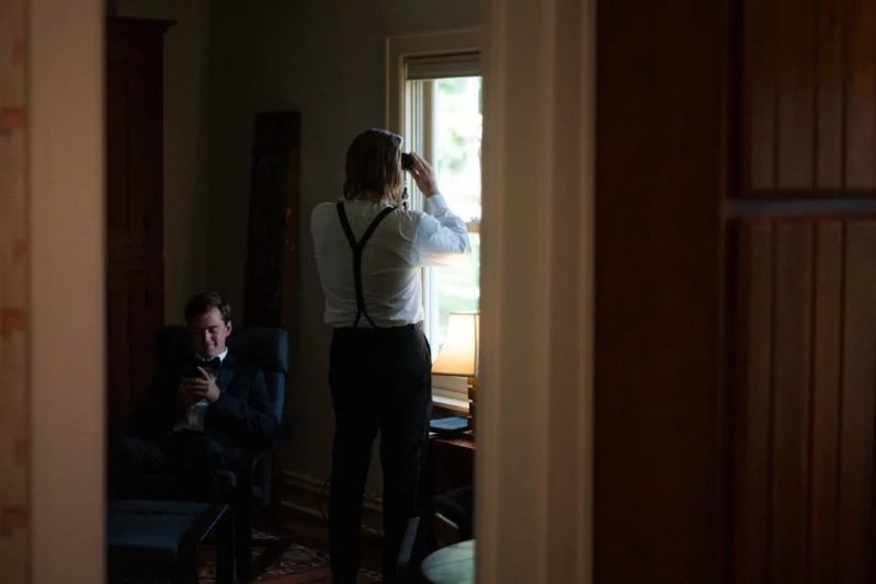 A woman and a man in a cozy room with wooden walls; the woman looking out the window and the man sitting in a chair using his phone.