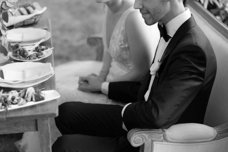 A black-and-white photo of a bride and groom sitting at a table during their wedding reception. The groom is dressed in a tuxedo, and the bride is wearing a lace wedding gown. They are seated side by side, next to a table with food.