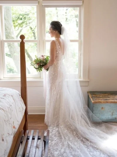 Bride in a white lace wedding gown holding a bouquet, standing by a window, in a bedroom.